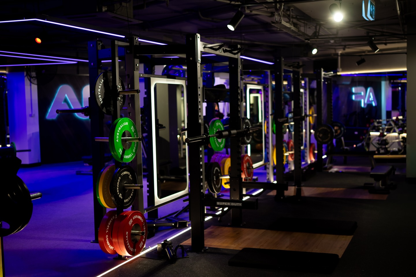 Wide view of a gym at night with neon signage and racks of colourful plates